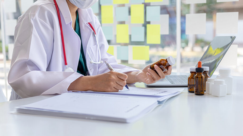 白衣を着た医療従事者が、処方薬の瓶を確認しながら薬歴や書類を作成している手元の様子